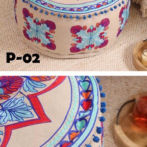 May include: A round, beige ottoman with blue pom-poms around the edge. The top of the ottoman is decorated with colorful embroidered flowers and geometric patterns.