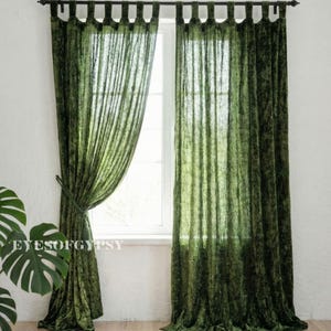Puede incluir: Cortinas de terciopelo verde colgadas de una barra oscura, enmarcando una ventana. Las cortinas están atadas en el lado izquierdo, revelando la ventana y un vistazo al exterior. La tela tiene una apariencia texturizada.