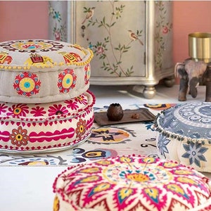 May include: Three round, colorful, embroidered floor cushions with a floral and geometric design. The cushions are stacked on top of each other, with the top cushion being a light gray color with a darker gray floral design.
