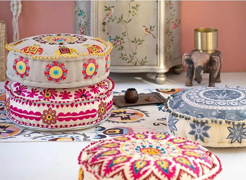 May include: Three round, colorful, embroidered floor cushions with a floral and geometric design. The cushions are stacked on top of each other, with the top cushion being a light gray color with a darker gray floral design.