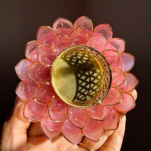 May include: A lotus flower candle holder featuring translucent pink petals with gold-toned wire edges. The center consists of a circular gold-toned metal cup with a decorative lattice pattern along the rim, designed to hold a small tea light candle.