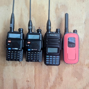 May include: Four handheld two-way radios with aerials are displayed against a wood grain background. Three are black, and one is coral-coloured. Each radio has a screen and a keypad. The radios are likely used for communication.