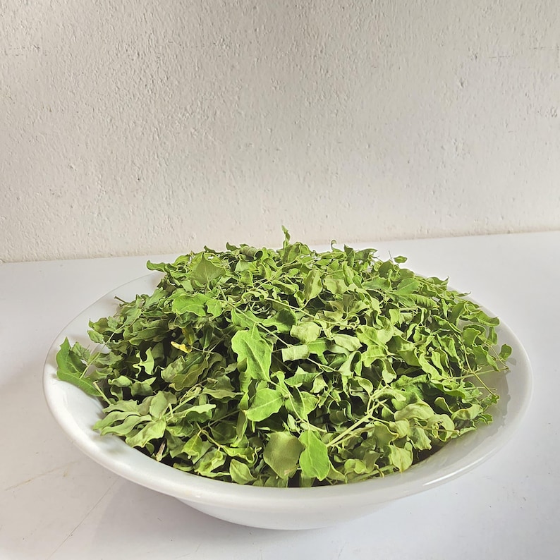 May include: A white, shallow bowl filled with dried green leaves. The leaves are a vibrant green color and appear to be a type of herb or spice. The bowl is set against a plain white background.