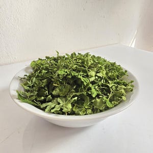 May include: A white ceramic bowl filled with dried green leaves. The leaves are small and have a slightly curled appearance. The bowl is sitting on a white surface, and the background is a plain white wall.