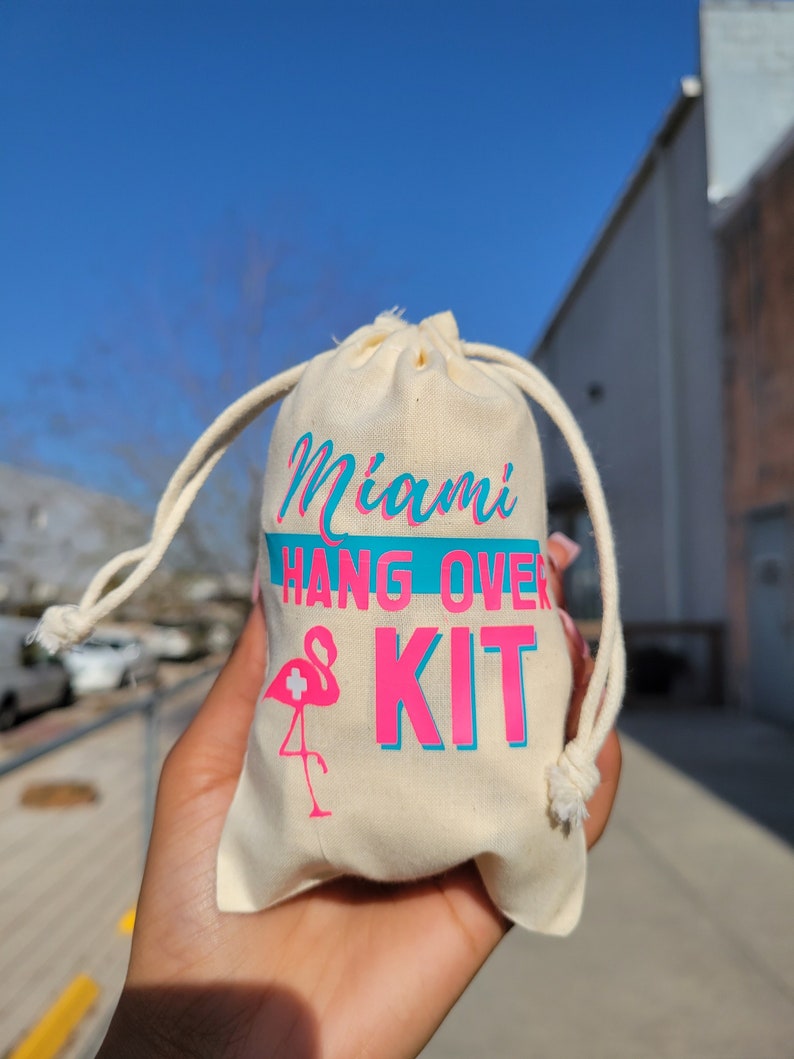 Puede incluir: Una bolsa de cord&oacute;n blanco con el texto "Miami Hangover Kit" en letras rosa y azul. Un flamenco rosa con un signo m&aacute;s blanco est&aacute; impreso en la bolsa.