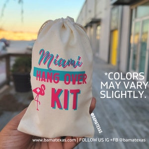 Puede incluir: Una bolsa de tela blanca con el texto "Miami Hang Over Kit" impreso en letras azules y rosas. Un flamenco rosa con una cruz blanca est&aacute; impreso en la bolsa. El texto "*COLORS MAY VARY SLIGHTLY." est&aacute; impreso debajo del nombre del kit. La bolsa est&aacute; sostenida en una mano.