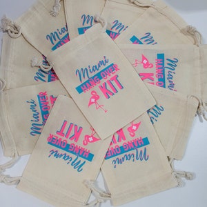 Puede incluir: Un grupo de diez peque&ntilde;as bolsas de algod&oacute;n blancas con cord&oacute;n de ajuste con texto rosa y azul que dice "Miami Hangover Kit". Las bolsas est&aacute;n apiladas unas encima de otras.