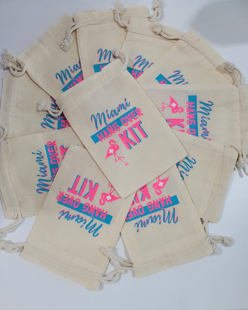 Puede incluir: Un grupo de diez peque&ntilde;as bolsas de algod&oacute;n blancas con cord&oacute;n de ajuste con texto rosa y azul que dice "Miami Hangover Kit". Las bolsas est&aacute;n apiladas unas encima de otras.
