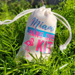 Puede incluir: Una bolsa de tela blanca con el texto "Miami Hangover Kit" en letras rosa y azul. Un flamenco rosa con una cruz blanca est&aacute; impreso en la bolsa.