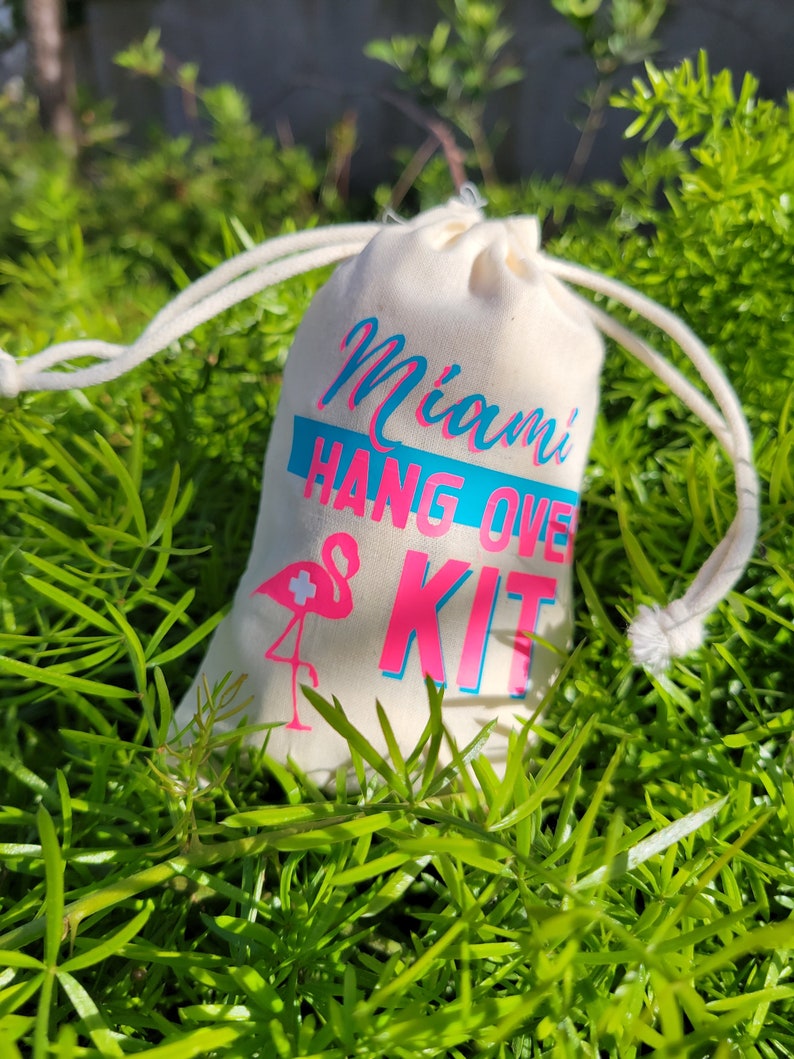 Puede incluir: Una bolsa de tela blanca con el texto "Miami Hangover Kit" en letras rosa y azul. Un flamenco rosa con una cruz blanca est&aacute; impreso en la bolsa.