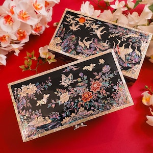 May include: Two rectangular black boxes with intricate floral and bird designs, accented with iridescent mother-of-pearl. The boxes are set against a vibrant red background, adorned with white and orange orchids.