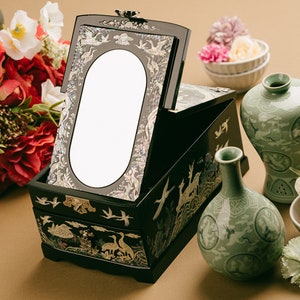 A black lacquer jewelry box with mother-of-pearl inlay and mirror, set on a beige surface with a bouquet of flowers and traditional green ceramic vases.