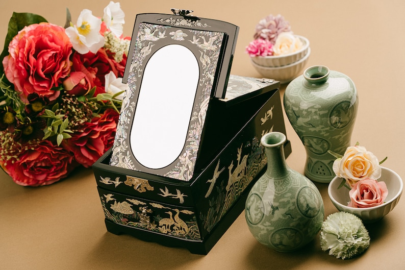 A black lacquer jewelry box with mother-of-pearl inlay and mirror, set on a beige surface with a bouquet of flowers and traditional green ceramic vases.