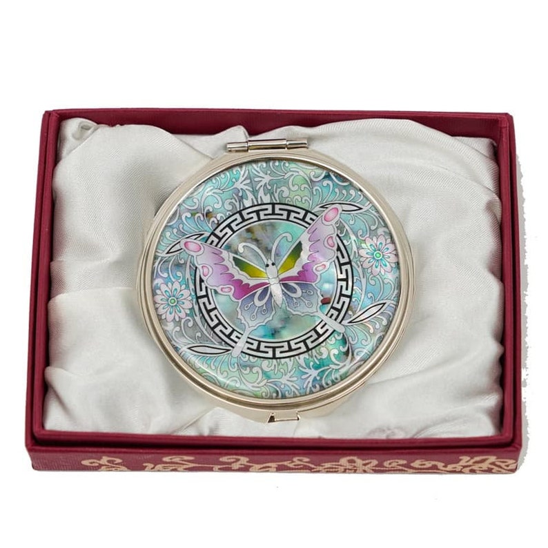 A compact mirror with a light blue, pink, and green butterfly design in a Greek key bordered circle, presented in a red box lined with white satin.