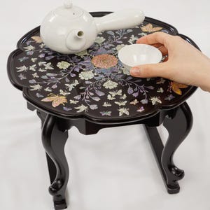 May include: A small, black, decorative table with a floral and butterfly design on the tabletop. A white teapot and a small white teacup are on the table. The table has curved legs.