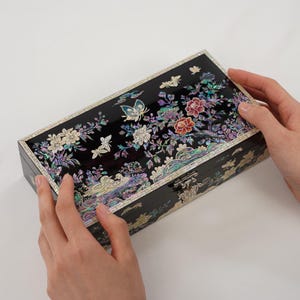 May include: A black rectangular jewelry box with a floral and butterfly design. The box is decorated with iridescent mother-of-pearl inlays, featuring white, pink, and purple flowers. It has a silver clasp and is held by two hands.