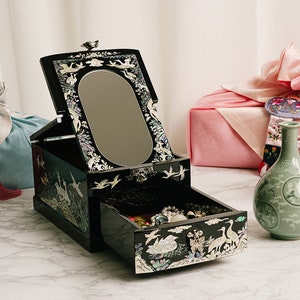 An open black jewelry box with intricate designs on a marble table, flanked by a blue pouch, a pink gift, and two green vases.