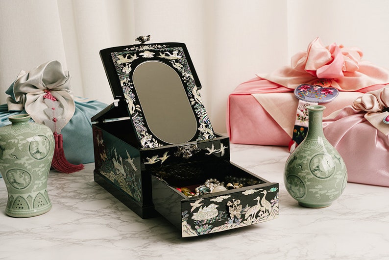 An open black jewelry box with intricate designs on a marble table, flanked by a blue pouch, a pink gift, and two green vases.