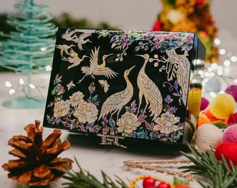 May include: Black lacquered box with mother-of-pearl inlay featuring cranes, flowers, and foliage. The box has a rectangular shape with a hinged lid and a silver-colored clasp. The design includes flying cranes and floral elements.