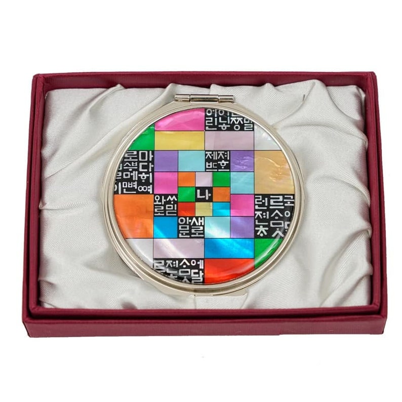 A round, colorful, mirrored compact with Hangul (Korean script) characters in various hues, presented in a red gift box with a white satin lining