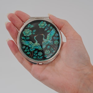 Peut inclure: Un miroir de poche rond et argenté avec un fond noir. Le miroir présente un motif de deux grues turquoise aux ailes déployées, entourées d'éléments floraux turquoise. Le miroir est tenu dans une main.