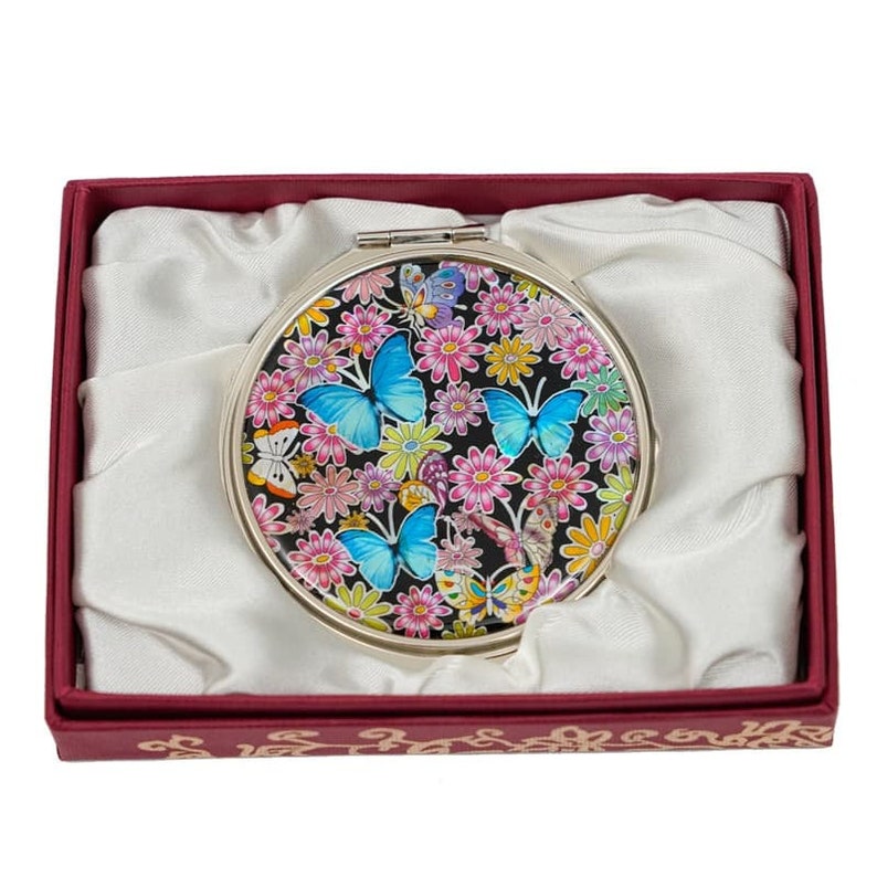 A compact mirror with vivid blue butterflies and colorful flowers on it, nestled in a red box with a cream satin interior.