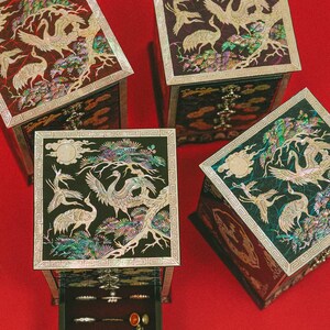 May include: Four decorative jewelry boxes with crane and tree designs. The boxes are black with inlaid iridescent accents. One box is open, revealing rings and a pendant. The boxes are on a red surface.