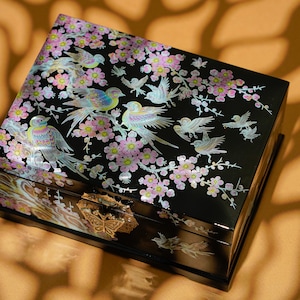 Könnte beinhalten: Eine schwarze Holzschatulle mit einem floralen und Vogeldesign, das mit Perlmutt eingelegt ist. Die Schatulle hat einen goldenen Schmetterlingsverschluss.