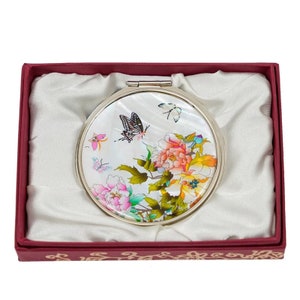 A round compact mirror with a floral and butterfly design, encased in a red box with a satin lining and decorative edging