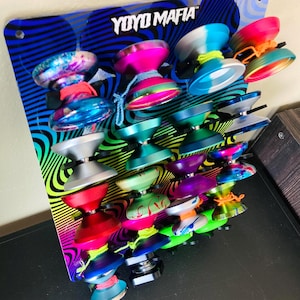 May include: A display of colorful yo-yos with the text "YoYo Mafia" on a patterned background. The yo-yos are in various colors, including pink, blue, green, and silver, with matching strings.