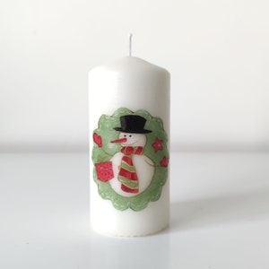 May include: White pillar candle with a snowman design. The snowman is wearing a black hat and a red and green scarf. The snowman is holding a red gift box. The candle is decorated with a green wreath.