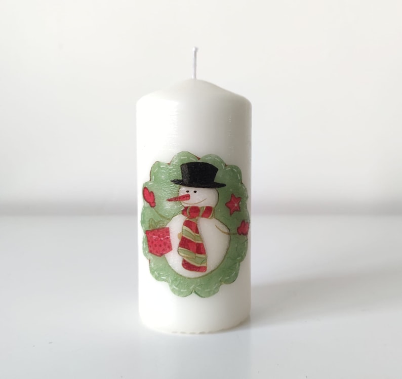 May include: White pillar candle with a snowman design. The snowman is wearing a black hat and a red and green scarf. The snowman is holding a red gift box. The candle is decorated with a green wreath.