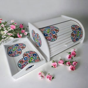 May include: White wooden bread bin and matching tray with a folk art heart design. The hearts are colourful with red, blue, green, yellow, and white. The bread bin has a hinged lid and a white handle.