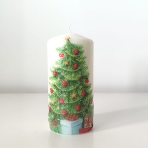May include: White pillar candle with a Christmas tree design. The tree is green with red ornaments and a gold star on top. There are presents at the base of the tree.