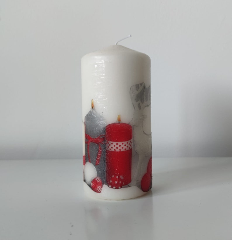 May include: White pillar candle with a Christmas design featuring red and gray candles, a reindeer, and red ornaments.