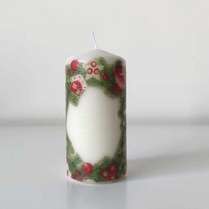 May include: A white pillar candle with a green and red Christmas design. The design features a wreath of pine branches with red berries and small houses.