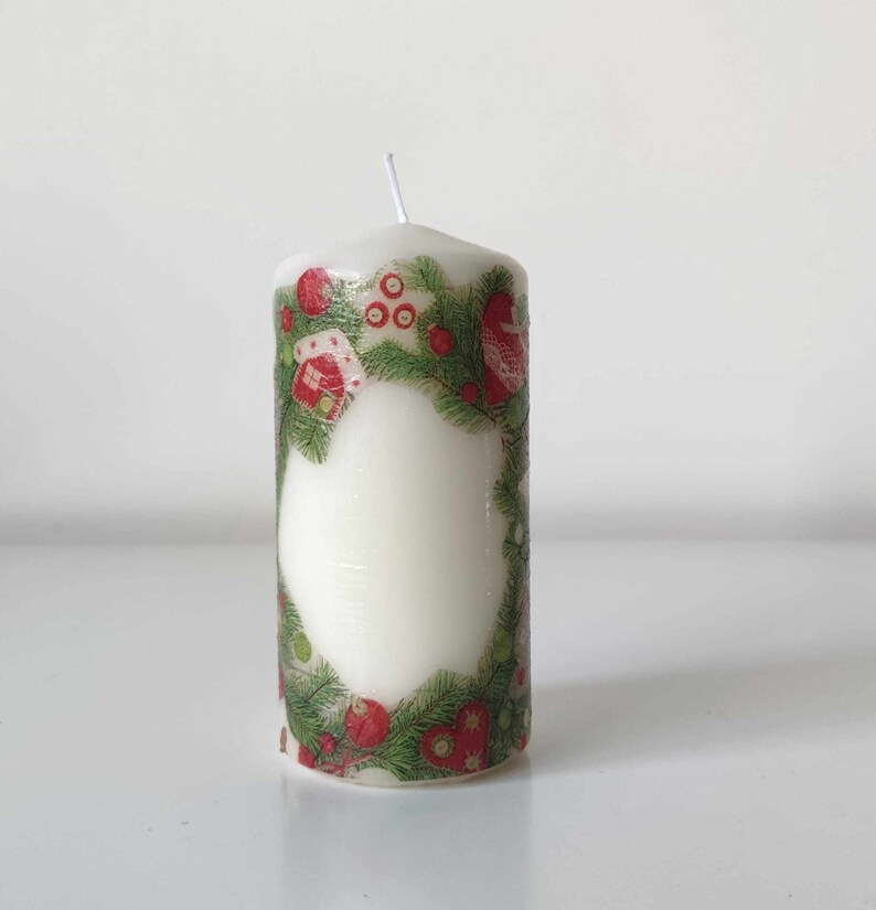 May include: A white pillar candle with a green and red Christmas design. The design features a wreath of pine branches with red berries and small houses.