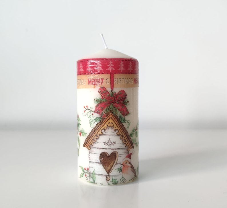 May include: White pillar candle with a Christmas design featuring a birdhouse, a red bow, and the text "Merry Christmas".
