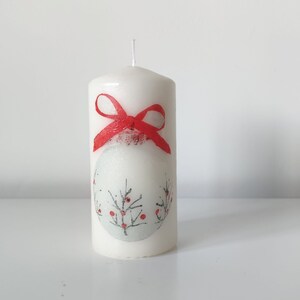 May include: White pillar candle with a red bow and a winter scene design. The design features a white ornament with a red berry branch.