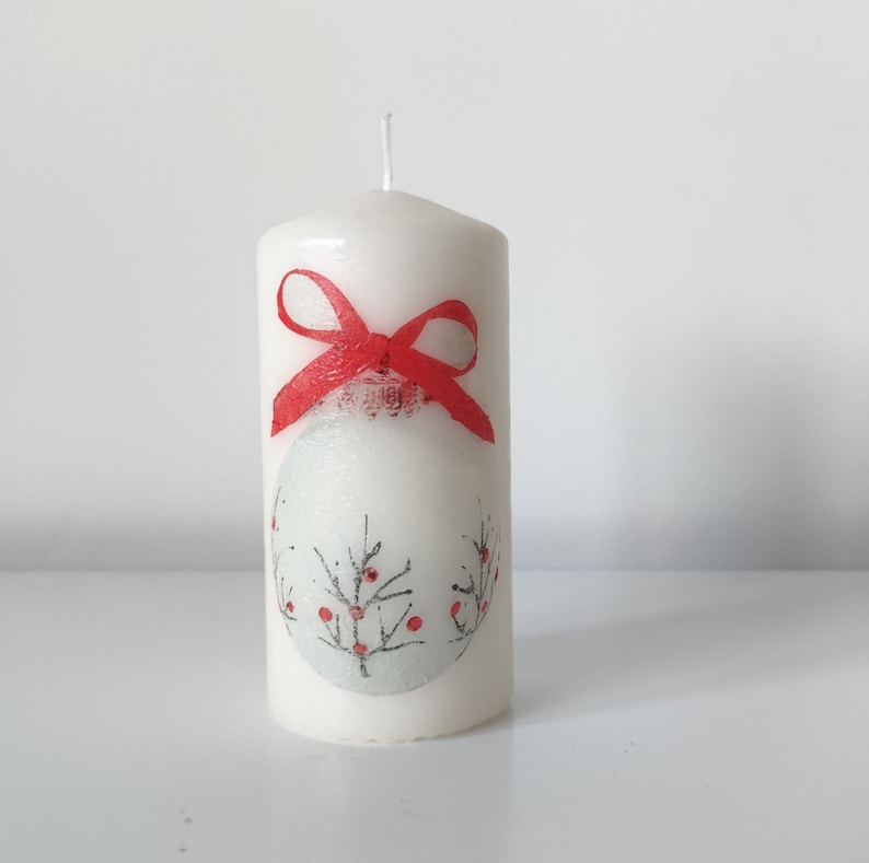 May include: White pillar candle with a red bow and a winter scene design. The design features a white ornament with a red berry branch.