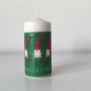 May include: White candle with a green and red design featuring three gnomes holding hands. The candle has the text "Merry Christmas" on the bottom.
