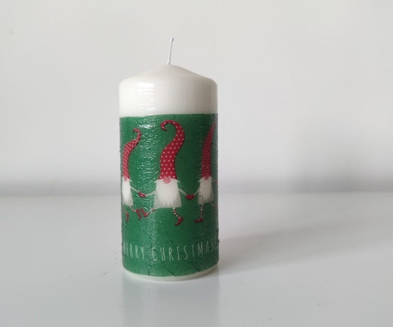 May include: White candle with a green and red design featuring three gnomes holding hands. The candle has the text "Merry Christmas" on the bottom.