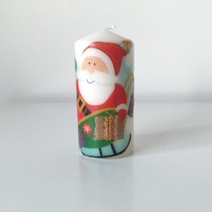May include: White candle with a colorful illustration of Santa Claus carrying a gift and riding a sleigh. The candle is decorated with a festive Christmas theme.
