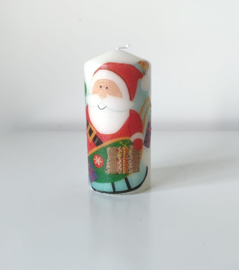 May include: White candle with a colorful illustration of Santa Claus carrying a gift and riding a sleigh. The candle is decorated with a festive Christmas theme.