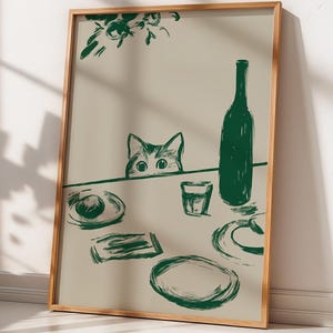 May include: A framed wall art print featuring a dark green line drawing on a light beige background. The illustration depicts a cat peeking over a table set with a wine bottle, a glass, plates, and a small rectangular object, with floral accents.