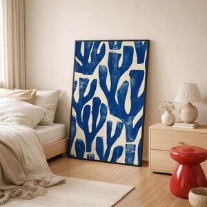 Blue abstract botanical poster in a white frame leaning against a white sofa, featuring deep navy leaf and coral-like organic shapes on a cream background, block print texture inspired by Matisse style cutout art. Modern coastal wall decor for living room or bedroom, ideal navy wall art print combining mid-century minimalist design with artisanal block printing texture — a striking indigo abstract statement piece for gallery walls and contemporary interiors.