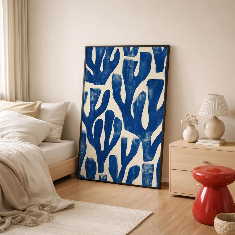 Blue abstract botanical poster in a white frame leaning against a white sofa, featuring deep navy leaf and coral-like organic shapes on a cream background, block print texture inspired by Matisse style cutout art. Modern coastal wall decor for living room or bedroom, ideal navy wall art print combining mid-century minimalist design with artisanal block printing texture — a striking indigo abstract statement piece for gallery walls and contemporary interiors.