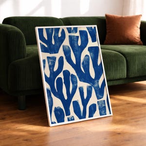 Blue abstract botanical poster in a white frame leaning against a white sofa, featuring deep navy leaf and coral-like organic shapes on a cream background, block print texture inspired by Matisse style cutout art. Modern coastal wall decor for living room or bedroom, ideal navy wall art print combining mid-century minimalist design with artisanal block printing texture — a striking indigo abstract statement piece for gallery walls and contemporary interiors.