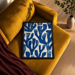 Blue abstract botanical poster in a white frame leaning against a white sofa, featuring deep navy leaf and coral-like organic shapes on a cream background, block print texture inspired by Matisse style cutout art. Modern coastal wall decor for living room or bedroom, ideal navy wall art print combining mid-century minimalist design with artisanal block printing texture — a striking indigo abstract statement piece for gallery walls and contemporary interiors.