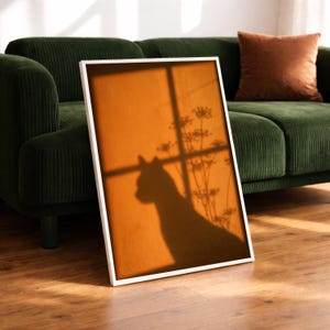 Cat shadow poster featuring a minimalist orange silhouette of a cat sitting by a sunlit window, with soft plant shadows on a textured amber wall. Moody aesthetic wall art perfect for bedrooms, reading nooks, or hallways. Warm sunset-toned cat silhouette print, a unique gift for cat lovers who enjoy dreamy, atmospheric wall decor rather than literal pet portraits.